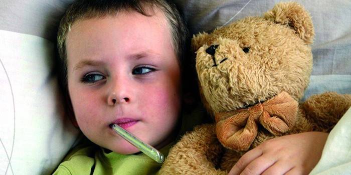 Un niño con un termómetro en la boca y con un oso de juguete en la mano.